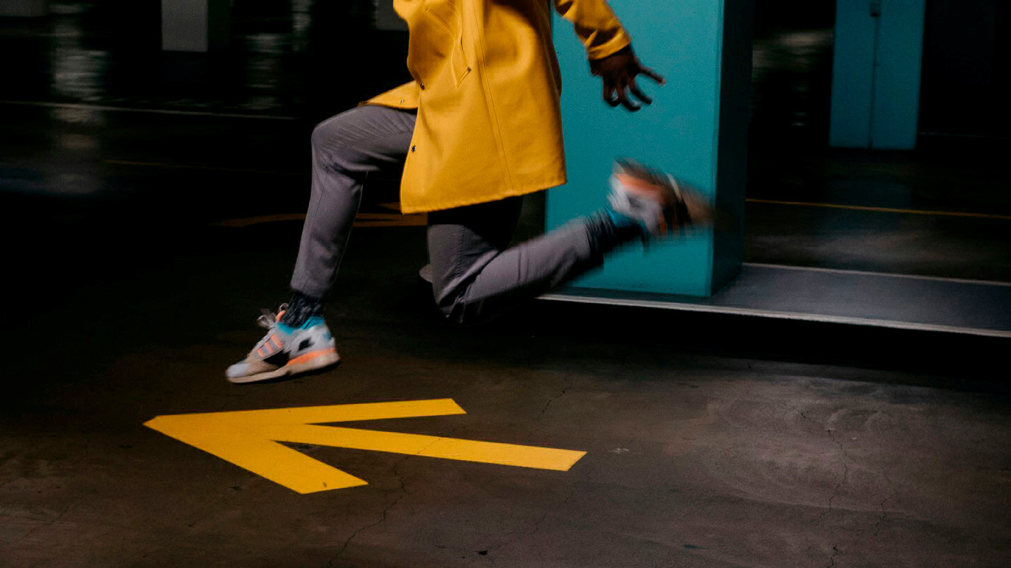 A person wearing a yellow raincoat jumps in the air over a yellow arrow on the ground.