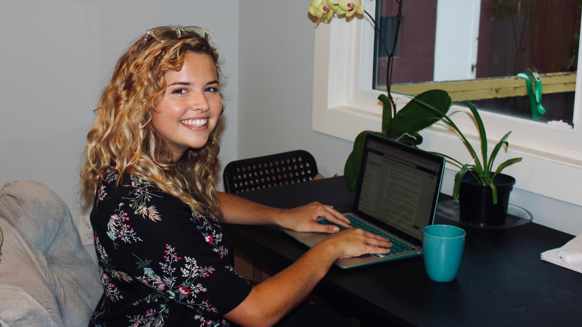 Co-op student Casey is smiling at the camera as she works on her laptop at a desk adorned with houseplants.