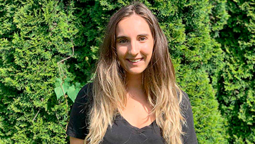 A student smiles while standing in front of a green tree.