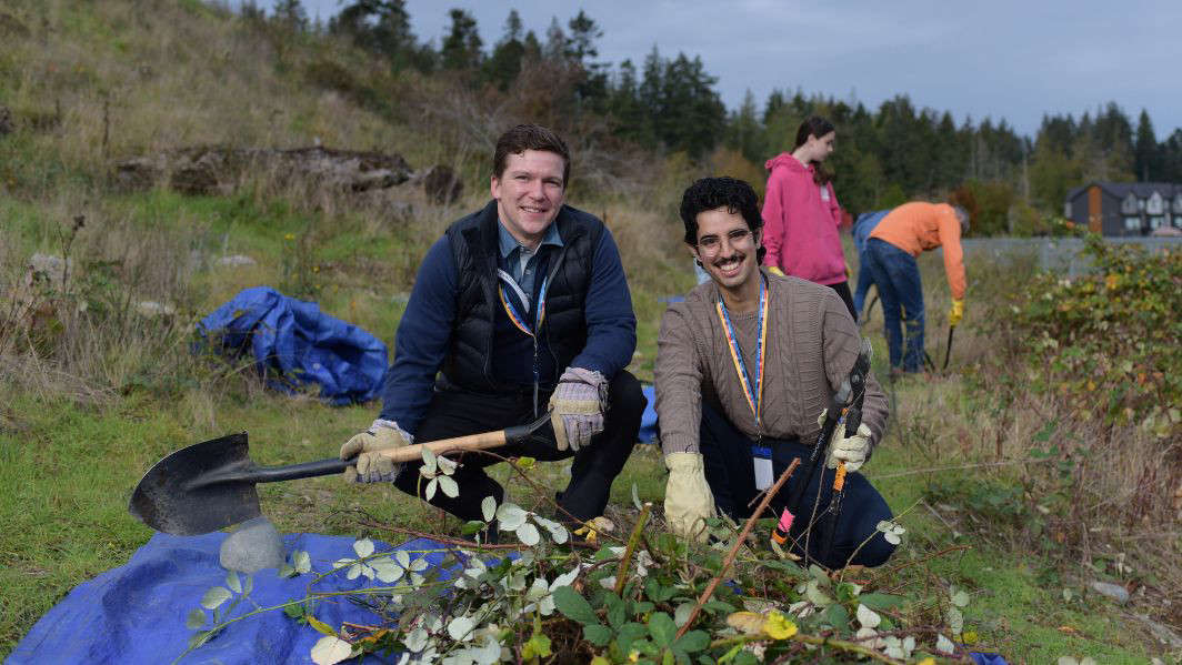 Two students are pictured working outdoors on practicum, clearing invasive blackberry bushes from a local school ground. They are crouched down side-by-side, smiling into the camera. One is holding a shovel and the other is holding large shears.