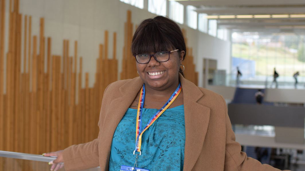 A student is pictured inside a school, with large glass windows in the background. She is wearing a blue shirt, glasses, and a UVic lanyard. She is smiling and looking into the camera.
