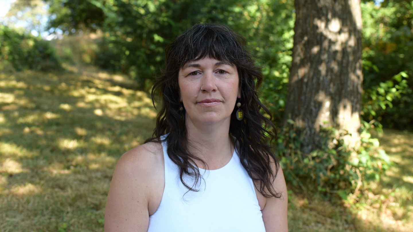 A head-and-shoulders portrait of Blythe Bell. She is pictured outdoors with a greenery background, and is looking into the camera lens. She is wearing a white tank top.