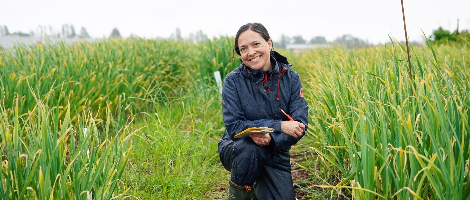 Brooke Hayes smiles at the camera, kneeling in a field of tall green grass holding a notebook and pen