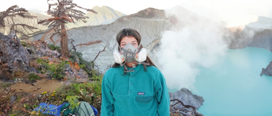 A woman in a gas mask stands before a mountain and lake