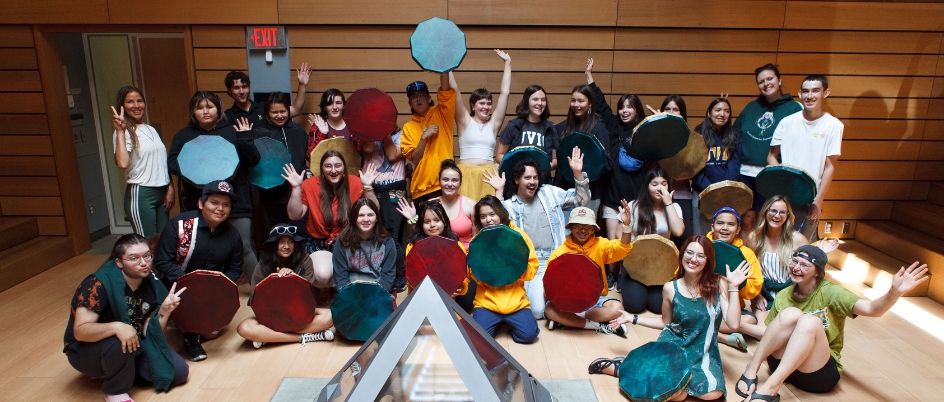 A large group of mini-U students and chaperones smile for the camera in the Ceremonial Hall; many are holding dyed hand drums and there's a grey indoor triangle fire pit in front of them