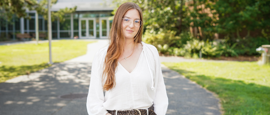 A student with long hair and glasses in a white blouse stands on the path leading to the UVic Bob Wright building