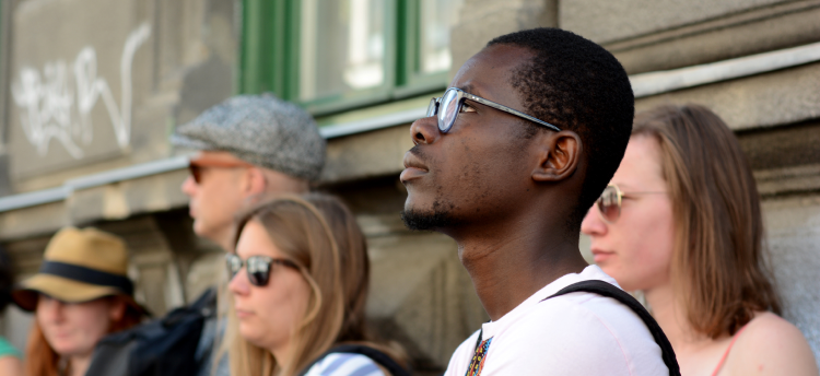 A group of students outside of a concrete building. A Black student with short hair, a beard, and glasses who is looking thoughtfully upwards is in focus in the foreground.
