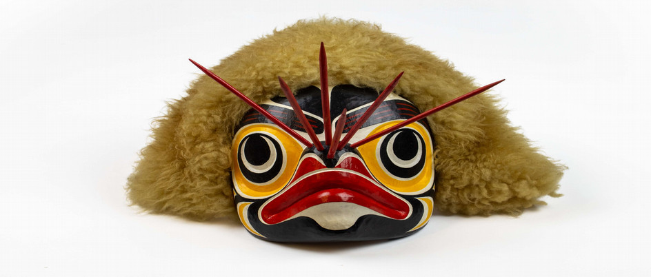 Small, brightly coloured mask depicting a bumblebee, resembling formline style. The mask is white with large black and red abstract designs, and two large black eyes. Four red stingers protrude from the area between the eyes. The mask is rimmed with fur.
