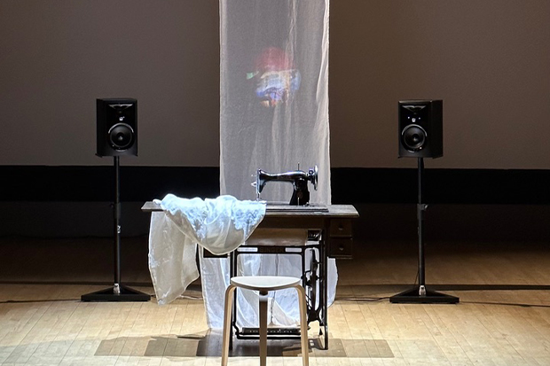 Dramatically lit photo of a vintage sewing machine holding a lenth of filmy cloth that stretches up to the ceiling, with an image projected onto the hanging cloth.