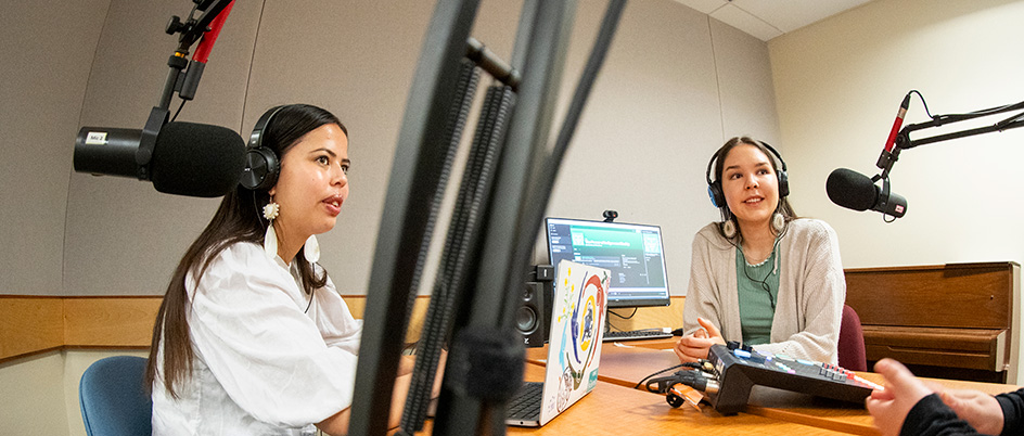 Two young Indigenous students sit at a desk with microphones overhead, wearing headphones.