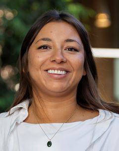 Smiling face of a woman with brown eyes and brown hair. She wears a white collared shirt over a white t-shirt.